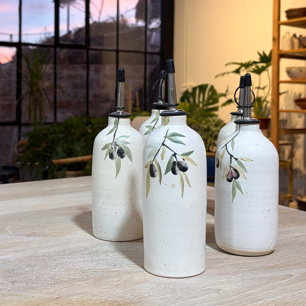Five white ceramic olive oil jugs with painted olive branches and metal pour spouts arranged on a wooden table.